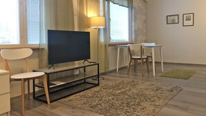 A modern living room with a flat-screen TV on a stand, a dining table with chairs, a lamp, and a large window with curtains. - Muurame-City Studios (Muurame)