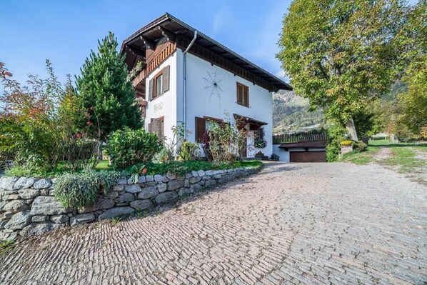 Une maison à deux étages avec un mur en pierre, une allée pavée et une montagne en arrière-plan.