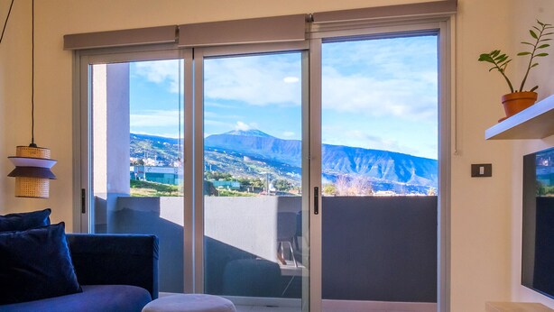 Un salón moderno con sofá, reposapiés y mesa de vidrio. Cuenta con un amplio ventanal corredero que da a un balcón con vistas a la montaña.
