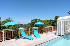 Un espace aménagé au bord de la piscine, avec deux parasols bleus, des chaises longues et une vue sur le paysage.