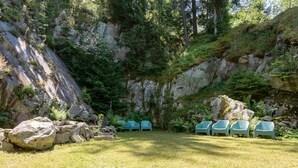 A grassy area with a row of green plastic chairs, a rocky cliff, and dense greenery. - Chalet 'Chalet Les Moraines 8' with Mountain View, Shared Pool and Private Terrace (Le Tholy)