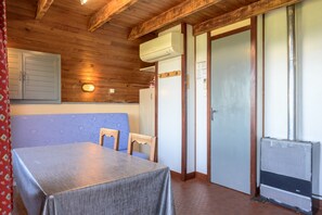 Une pièce avec un plafond en bois, une table recouverte d’une nappe, un banc bleu avec coussin, une porte blanche et un appareil de climatisation.