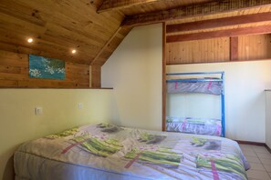Une chambre avec un lit superposé, un lit simple avec une literie à motifs floraux, des poutres en bois au plafond et un tableau accroché au mur.