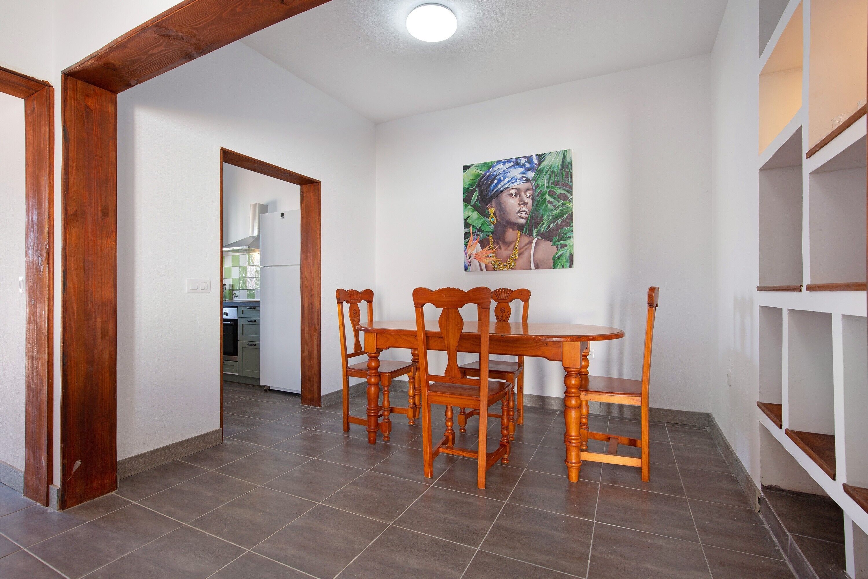A dining area with a wooden table and chairs, a painting on the wall, and a kitchen in the background.