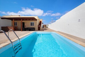 A swimming pool with a metal ladder, a covered patio, and a white building with a door and window.