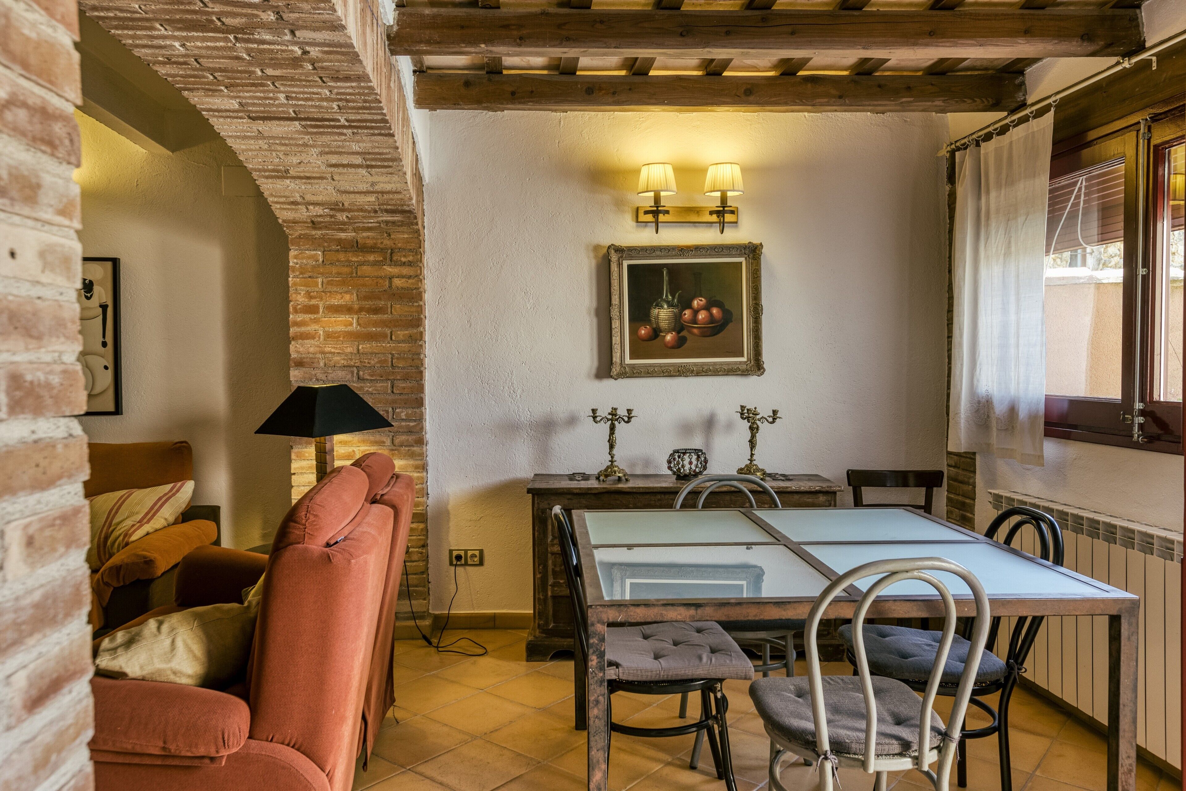 A cozy room with a brick wall, a wooden ceiling, a dining table with chairs, a sofa, and a lamp.