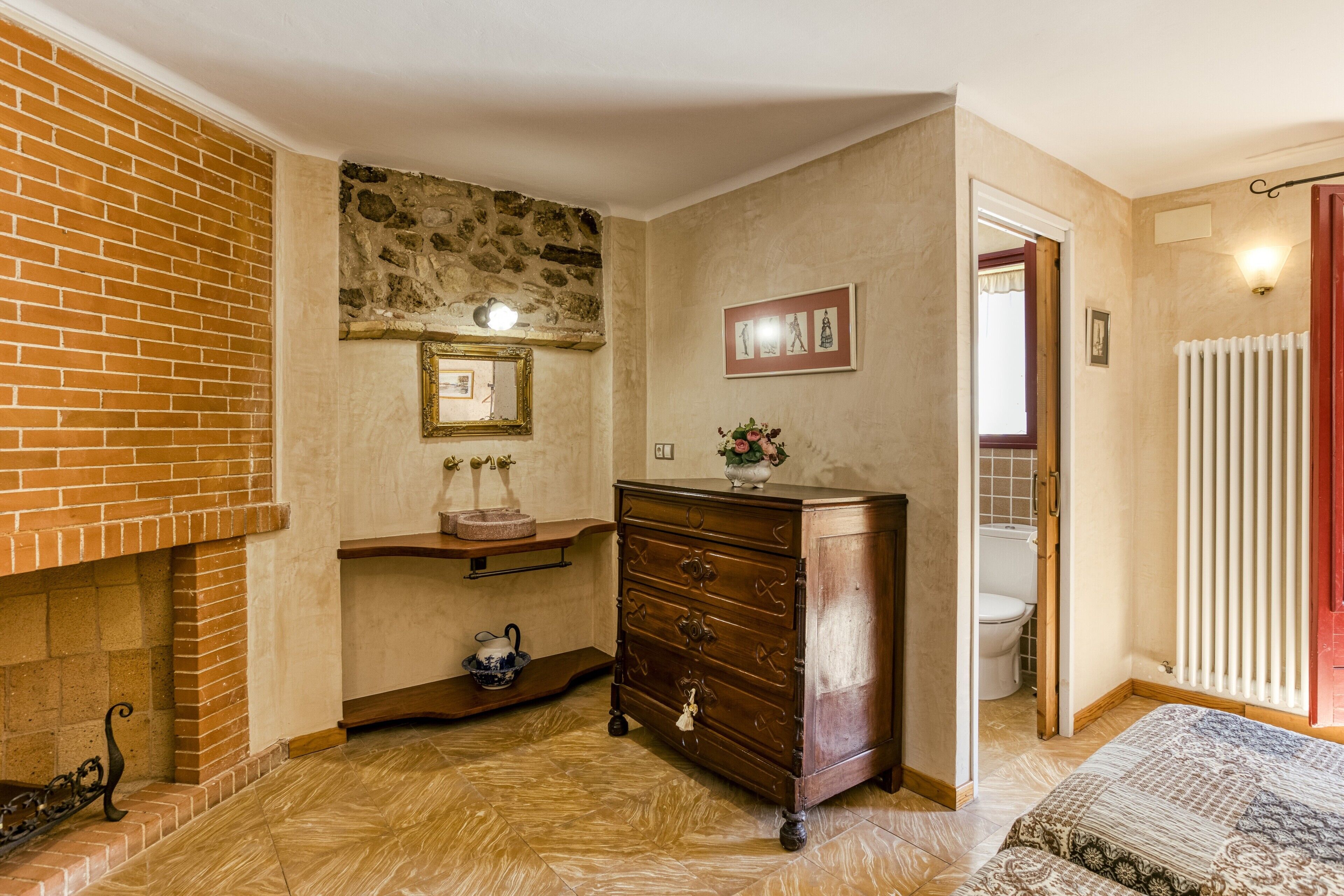 A room with a brick fireplace, a wooden dresser, a tiled floor, a framed picture on the wall, and a doorway leading to a bathroom.