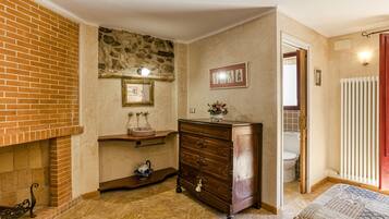 A room with a brick fireplace, a wooden dresser, a tiled floor, a framed picture on the wall, and a doorway leading to a bathroom.