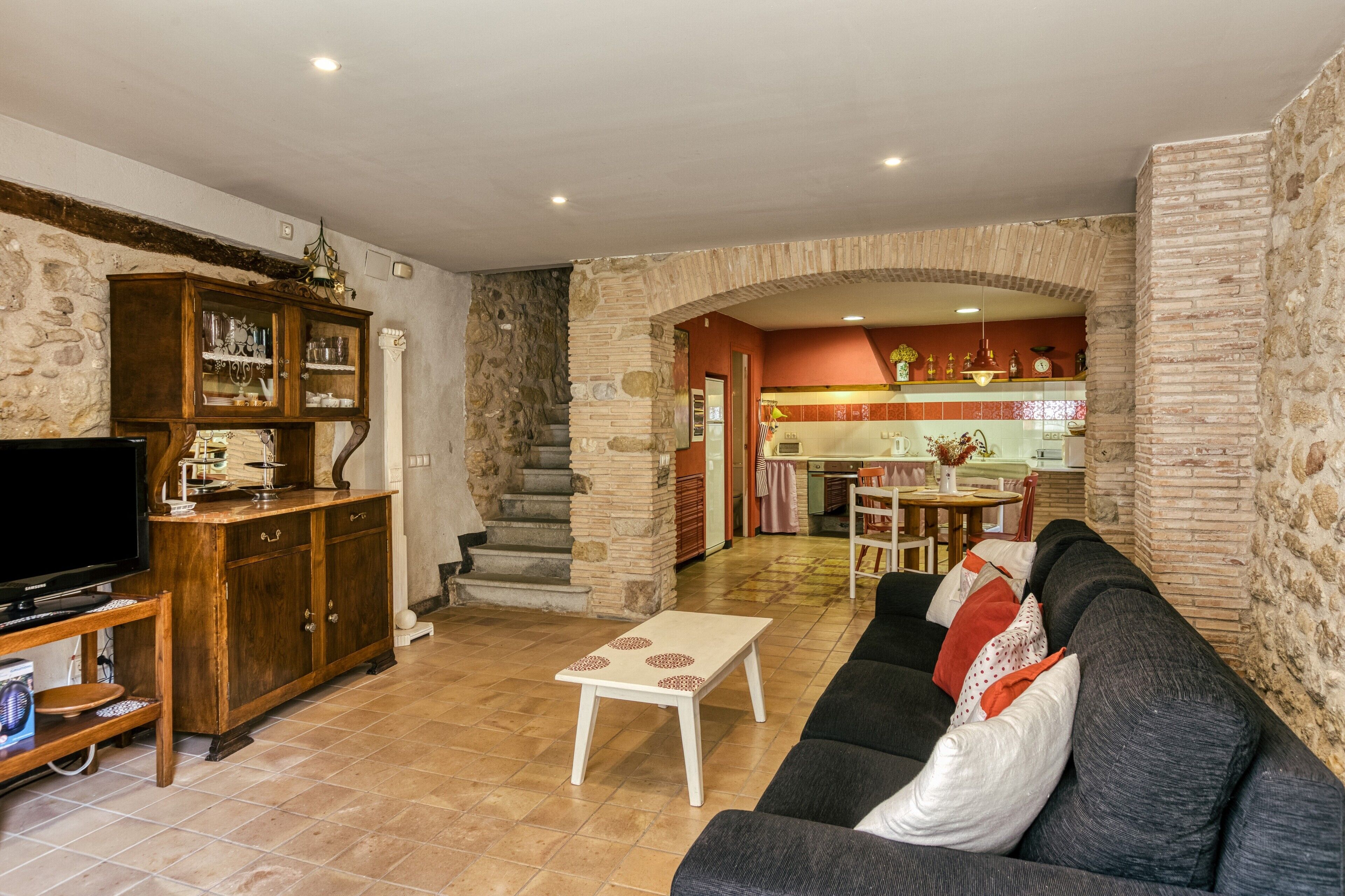 A living room with a sofa, TV, and wooden cabinet. There is a staircase leading to a kitchen area.