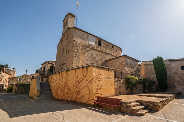 A stone building with a staircase leading to a door, a bench, and a flag on top.