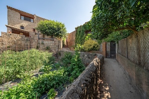 A narrow stone pathway flanked by lush greenery and a rustic stone building with a tiled roof.