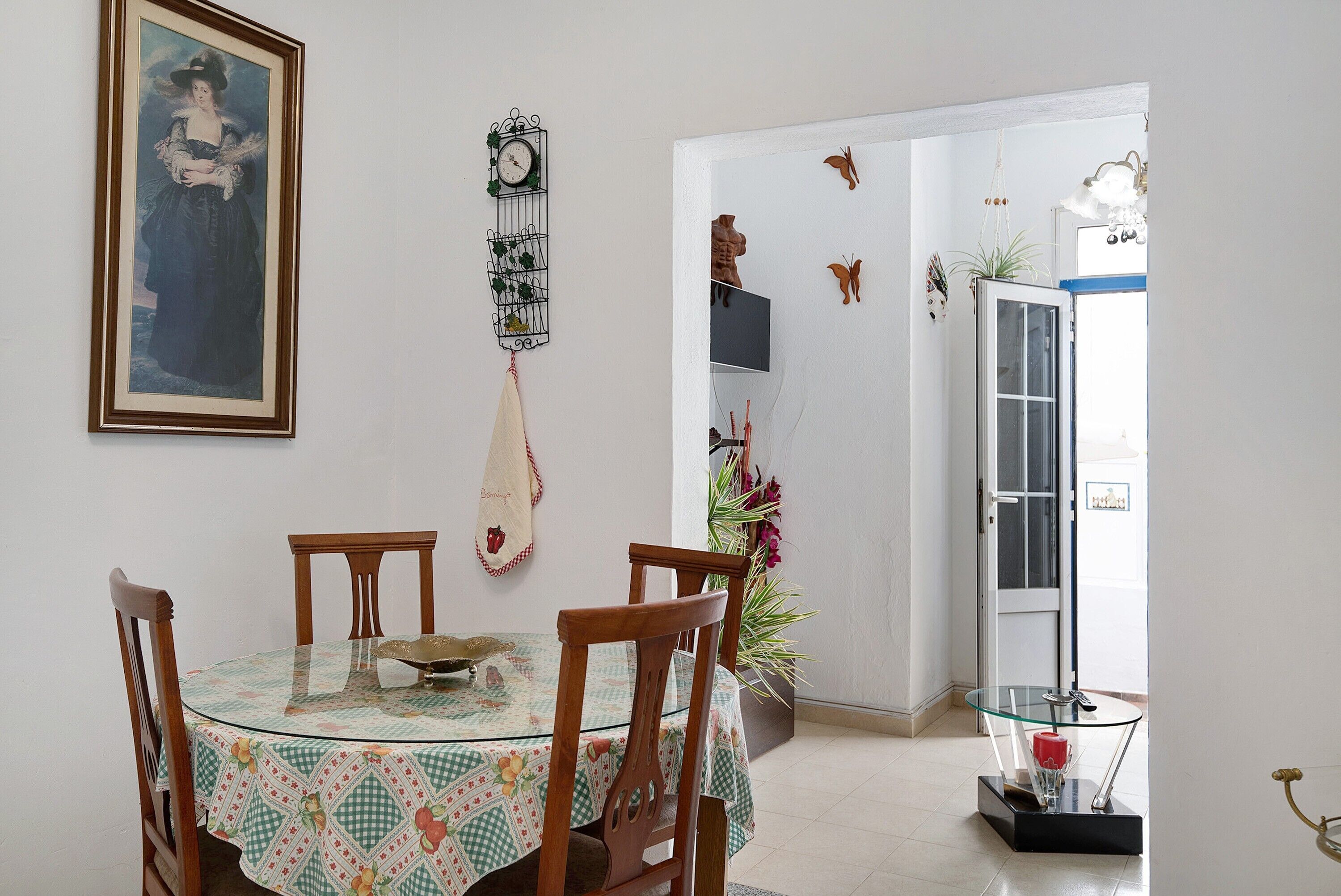 Un área de comedor con una mesa redonda de cristal, sillas de madera, un cuadro enmarcado, un reloj de pared y una puerta decorada con motivos de mariposas.