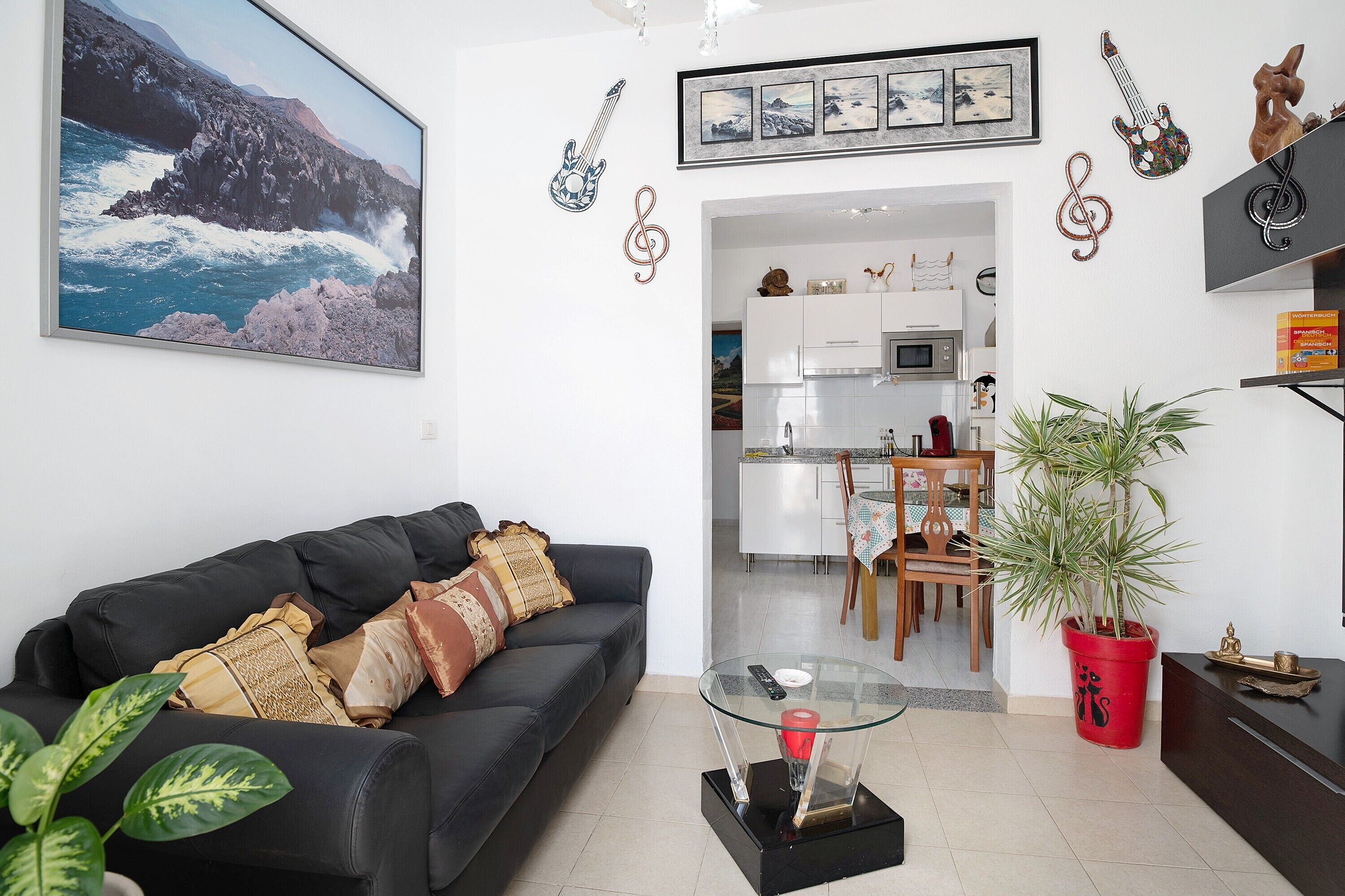 Una sala de estar con un sofá negro, una mesa de centro de vidrio, una planta en maceta roja y una cocina al fondo.