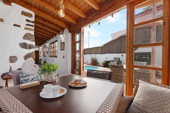 A cozy room with a wooden ceiling, stone wall, wicker furniture, a dining table with plates and cups, and a view of a swimming pool.