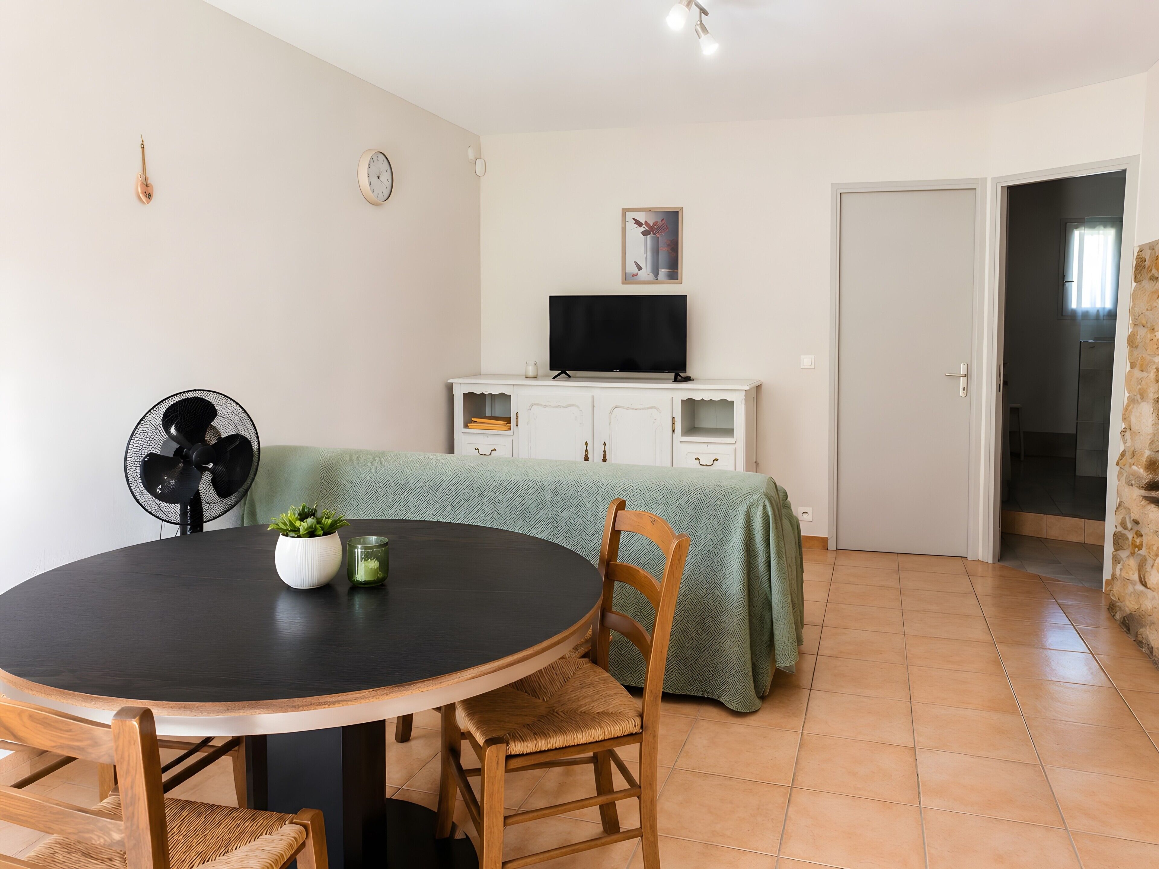 Un comedor con una mesa redonda, sillas, un ventilador, un televisor sobre un mueble y una puerta que da a otra habitación.