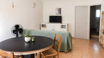 Uma sala de jantar com uma mesa redonda, cadeiras, um ventilador, uma televisão sobre um móvel e uma porta que dá para outro quarto.