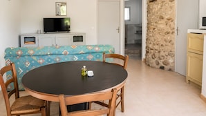 Un coin repas avec une table ronde et des chaises, un canapé recouvert d’un tissu à motifs, un téléviseur posé sur un meuble et un mur en pierre.