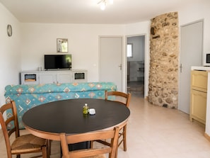 Un coin repas avec une table ronde et des chaises, un canapé recouvert d’un tissu à motifs, un téléviseur posé sur un meuble et un mur en pierre.