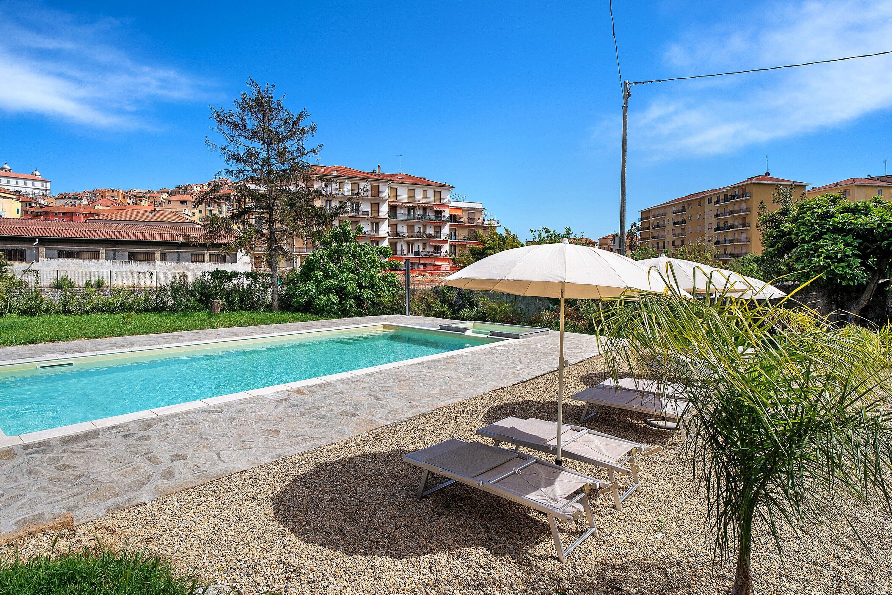 A swimming pool with lounge chairs and an umbrella, surrounded by pebbles and greenery, with buildings in the background.