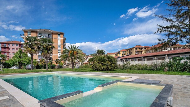 A swimming pool with a stone border, surrounded by buildings and greenery. - Holiday Home 'Il Gattino Di Porto Appartamento 1' with Shared Pool, Wi-Fi and Air Conditioning (Imperia)