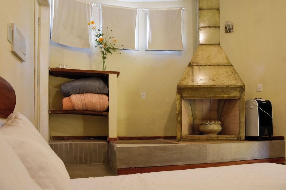 A room with a bed, a fireplace, a shelf with pillows, and a window with curtains.