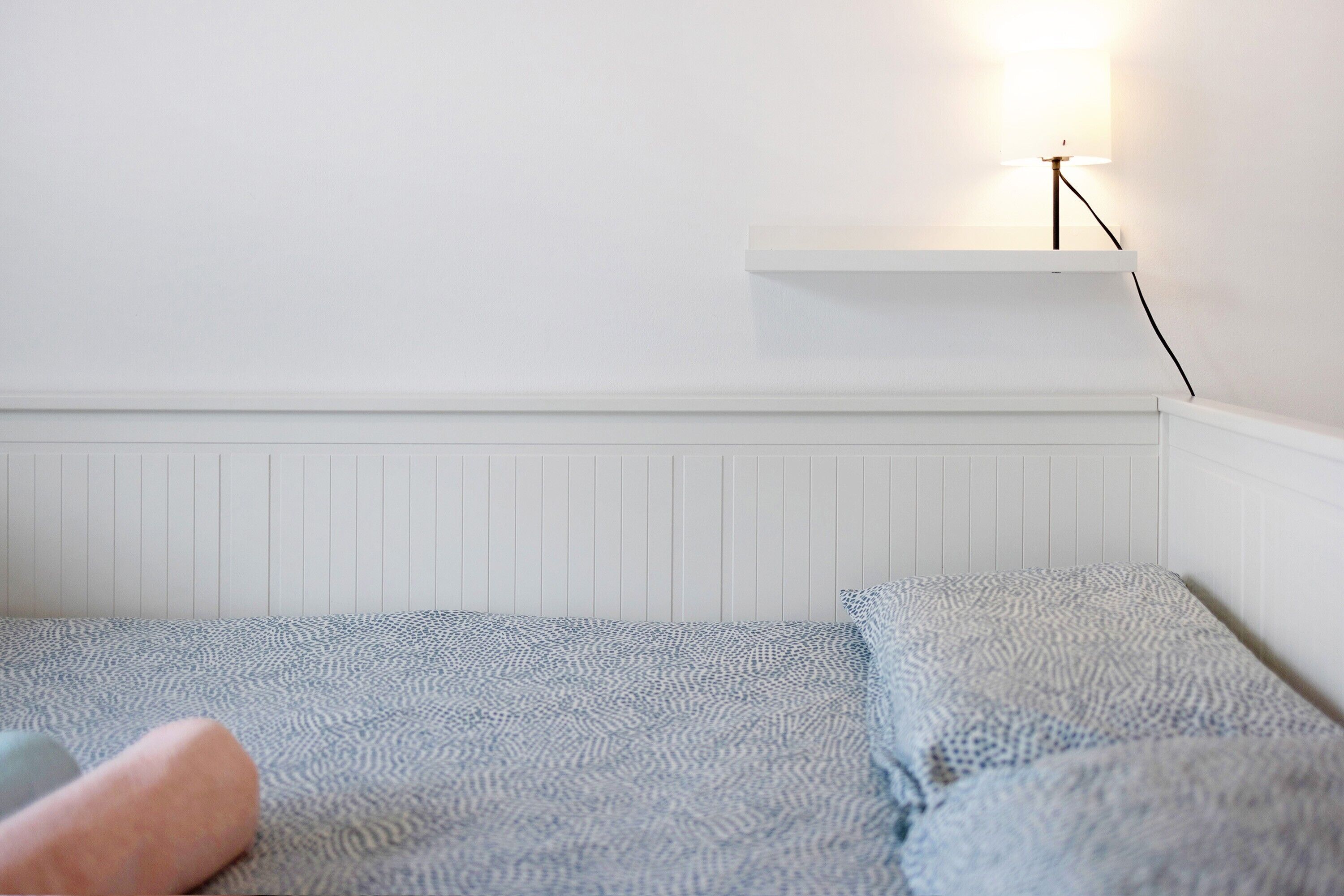 Een witte kamer met een bed, een witte plank en een wandlamp.