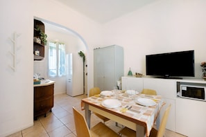 A dining area with a table set for two, a television on the wall, and a kitchen area with cabinets and a refrigerator.