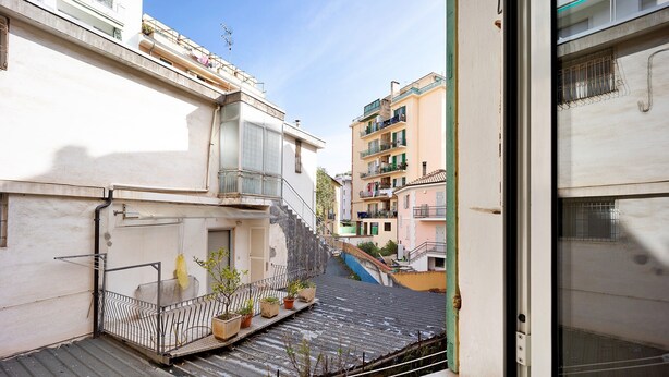 Een smalle stadstraat met gebouwen, een zichtbare trap en een balkon met plantenpotten.