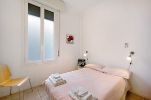 A bedroom with a bed, a chair, a window, a wall-mounted light, and a small picture.