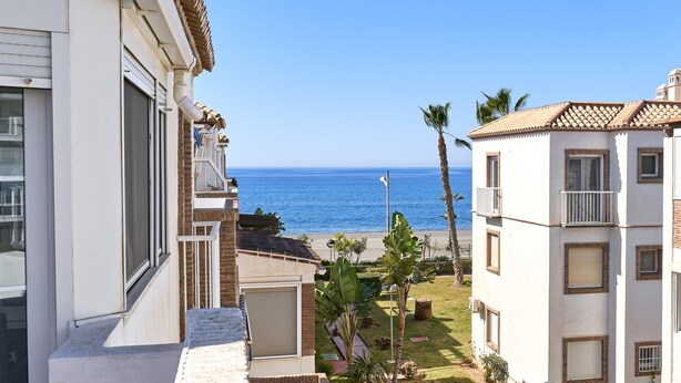 Um edifício costeiro com vista para o mar, uma varanda e um pequeno jardim com palmeiras.