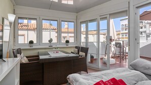 Quarto com cama, mesa de jantar com cadeiras, pequena cozinha e grandes janelas com vista para prédios.