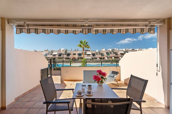 A balcony with a table, chairs, a vase of flowers, and a view of a swimming pool and buildings.