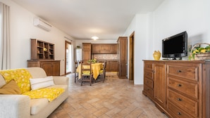 A living room with a sofa, a dining table with chairs, wooden cabinets, a television, and a kitchen area with a refrigerator. - Holiday Home 'Girasole' with Private Terrace, Garden and Air Conditioning (Pula)