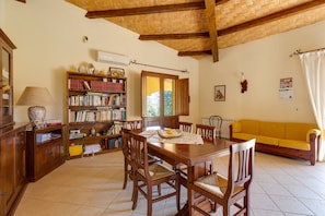 Un'area pranzo con un tavolo e sedie in legno, una libreria, un divano giallo e una finestra con vista su piante.