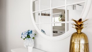 Een witte kaptafel met een ronde spiegel, een witte vaas met blauwe bloemen en een gouden vaas met een plant.