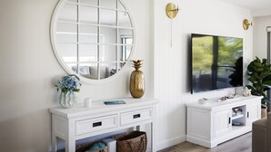 Een moderne woonkamer met een witte console tafel, een grote ronde spiegel, een flatscreen televisie en een potplant.