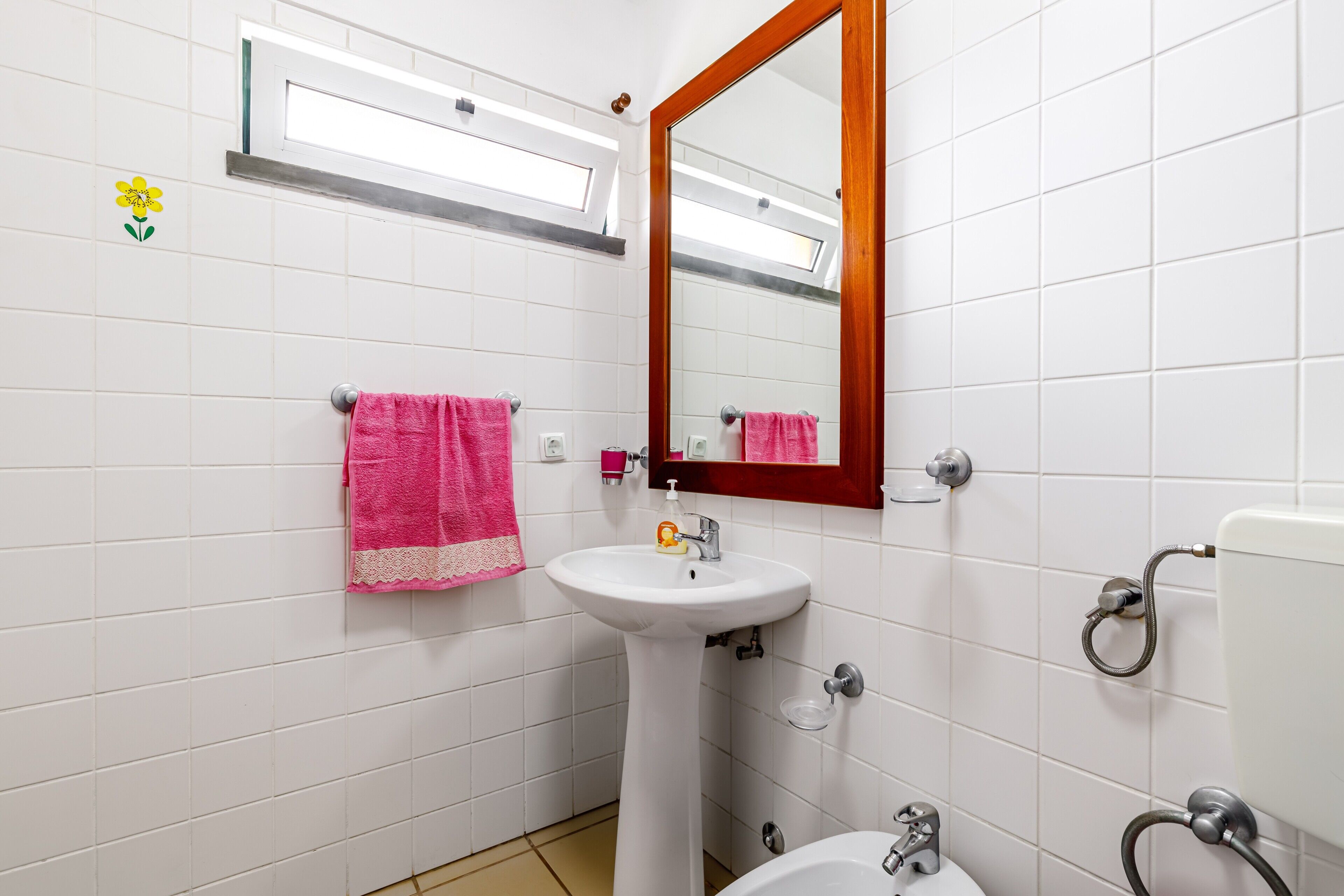Un baño con paredes de azulejos blancos, un lavamanos pedestal, un espejo con marco de madera y una ventana sobre el lavamanos.