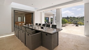 Ein moderner Essbereich mit großem Tisch und Korbstühlen, Blick auf einen angelegten Garten und ein geräumiges Interieur mit hohen Decken und hellem Tageslicht.