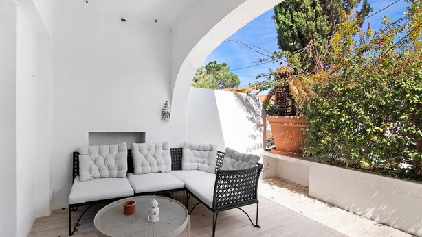 Un interior blanco con sofá y una mesa redonda, una pared blanca y vistas a árboles y una cerca.