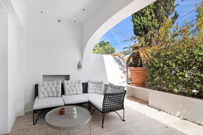 Um interior claro com sofá e mesa redonda, parede branca e vista para árvores e uma cerca.