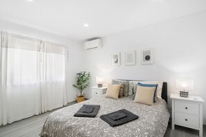 Un dormitorio con cama, mesitas de noche, aire acondicionado de pared, planta en maceta y ventana con cortinas.