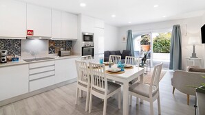 Una cocina moderna con armarios blancos, un comedor con sillas blancas y una sala de estar con sofá y televisor.