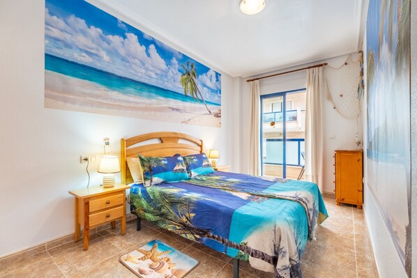 Une chambre à coucher avec un lit, des tables de chevet, une lampe, un tableau représentant une plage et une vue sur la mer par la fenêtre.