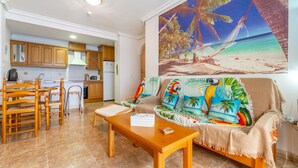 A living room with a wooden dining table, a sofa with a tropical print, and a kitchen in the background.