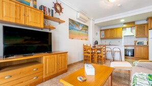 A living room with wooden furniture, a flat-screen TV, a dining area with chairs, and a kitchen with wooden cabinets.