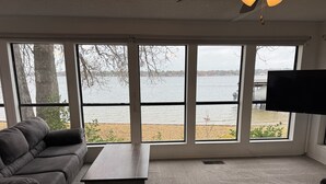 A living room with a large window overlooking a lake, a dark brown leather sofa, a wooden coffee table, and a flat-screen TV mounted on the wall. - Lakefront with Private Dock & Sandy Shore (Malakoff)