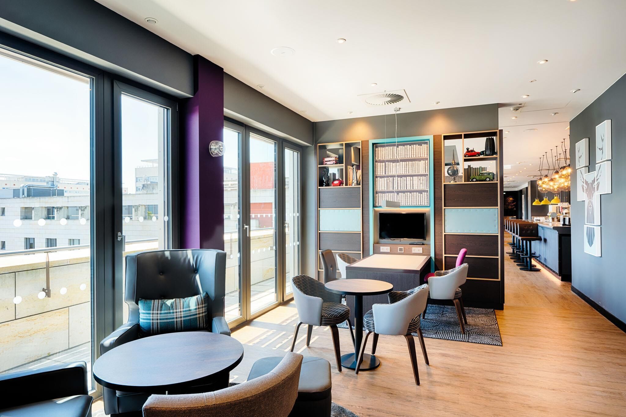 Un hall d’hôtel moderne doté de grandes fenêtres, d’un espace salon avec des tables et des chaises, d’une bibliothèque et d’un bar.