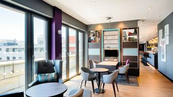Un hall d’hôtel moderne doté de grandes fenêtres, d’un espace salon avec des tables et des chaises, d’une bibliothèque et d’un bar.