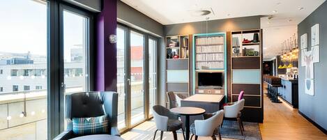 Un moderno vestíbulo de hotel con amplias ventanas, una zona de estar con mesas y sillas, una estantería y un bar.