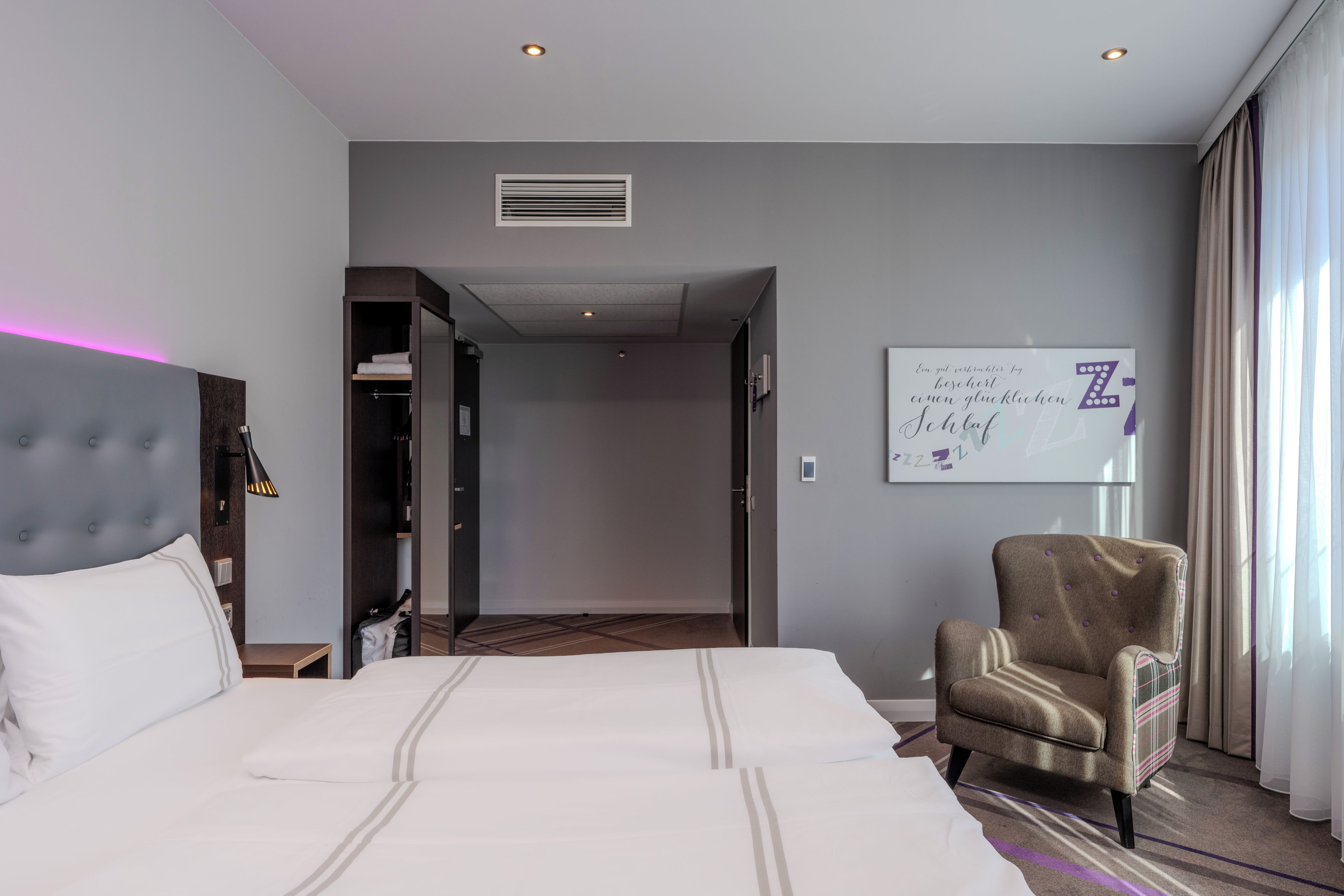 A hotel room with a bed, a chair, a wardrobe, and a wall with a sign.